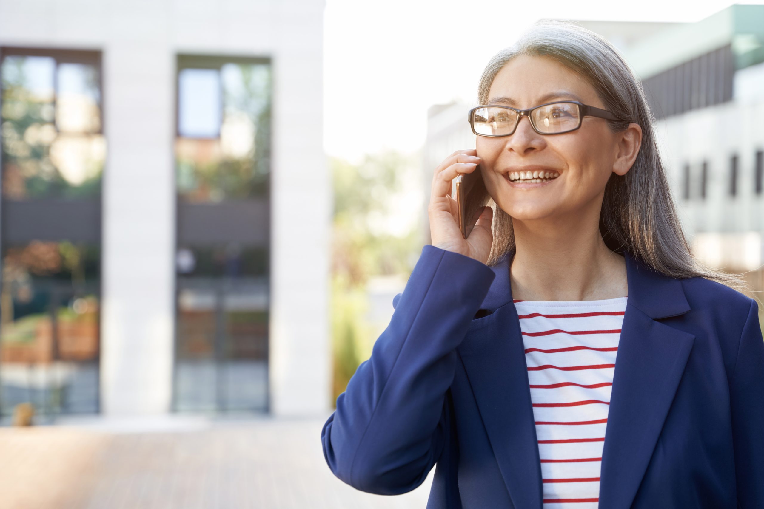 Good business talk. Portrait of cheerful mature business woman wearing eyeglasses and classic wear talking on mobile phone and smiling while standing against office building outdoors