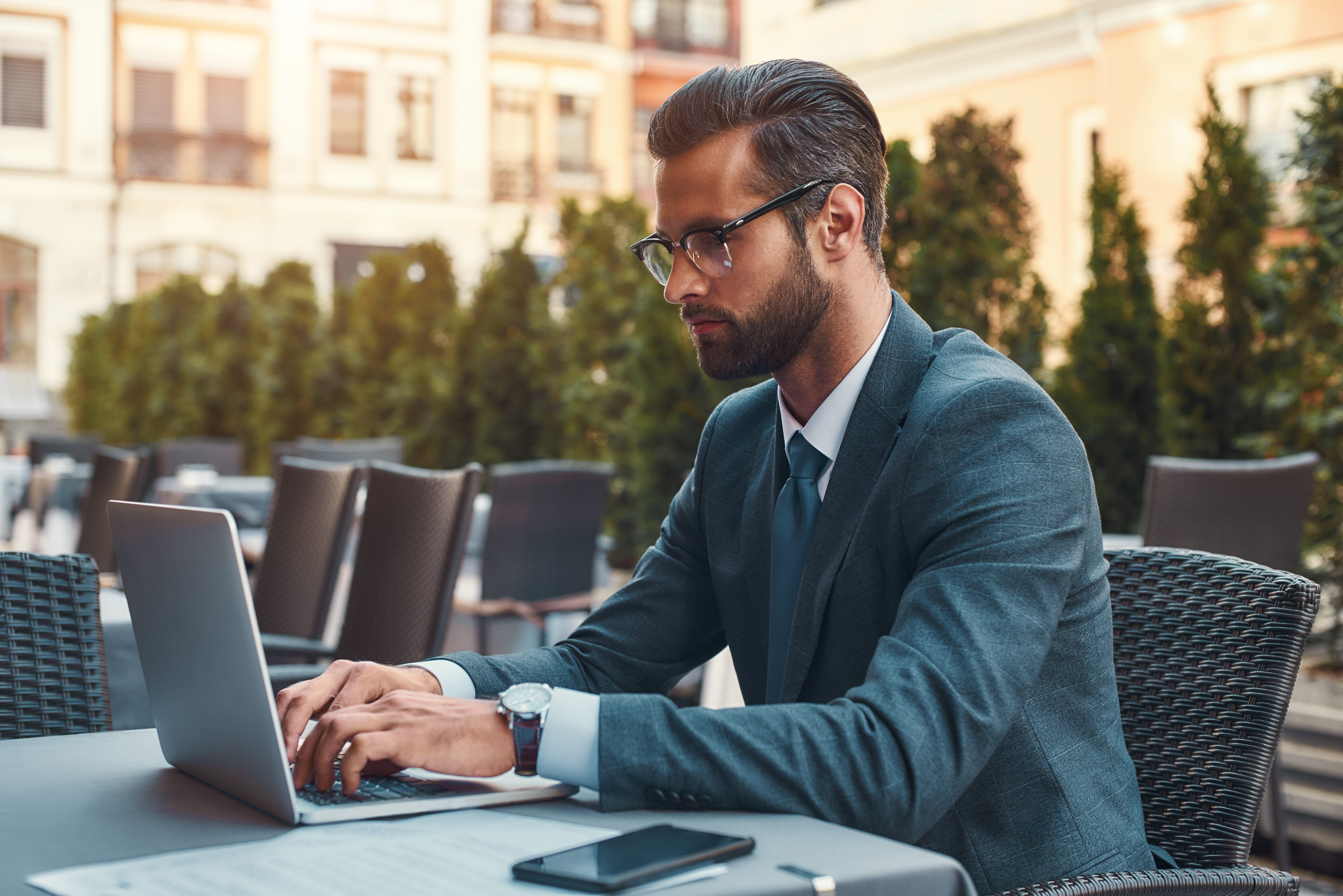 Modern businessman. Portrait of handsome bearded businessman in eyeglasses working with laptop while sitting in restaurant outdoors