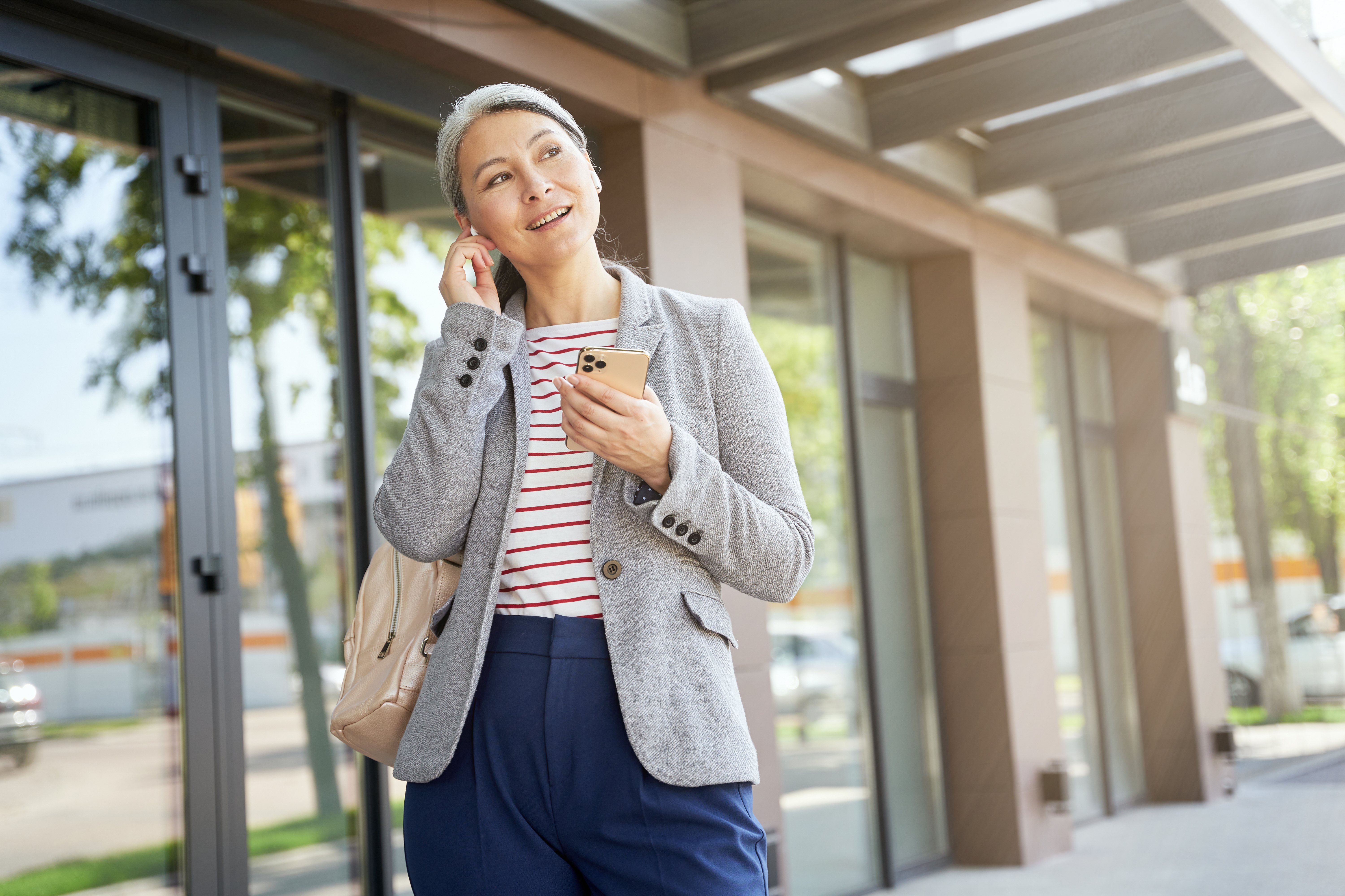 Portrait of a beautiful and happy woman wearing wireless earphones using smartphone, talking with somebody by phone while standing outdoors