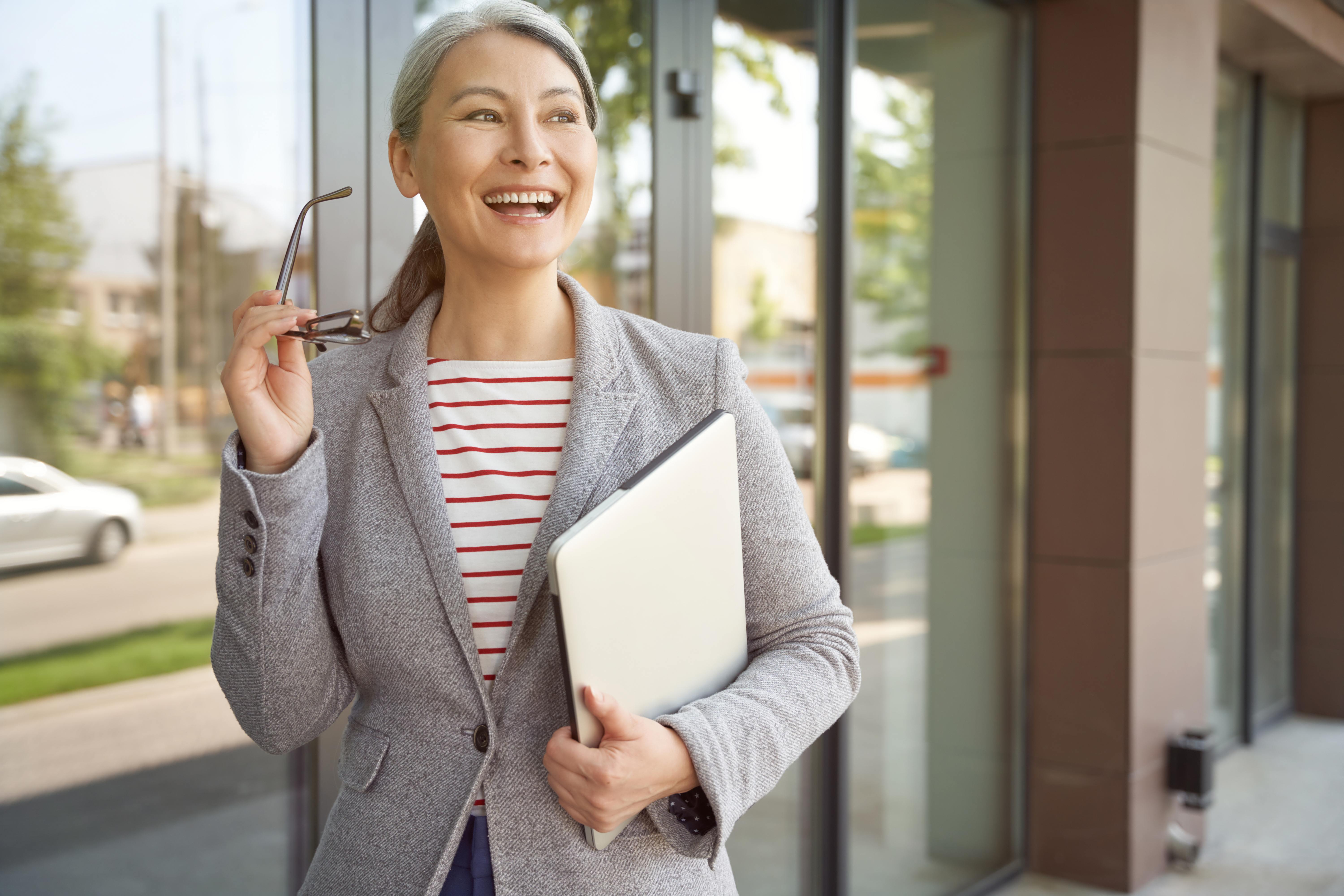 Ready to work. Beautiful and happy mature business woman holding laptop and smiling while standing near office building